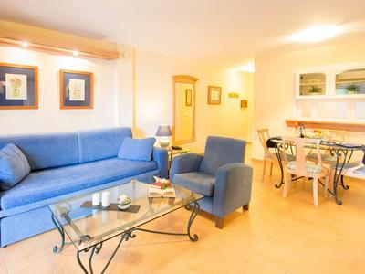 Helles Wohnzimmer mit blauem Sofa, Sessel, Glastisch und Esstisch in einem Hotelzimmer.