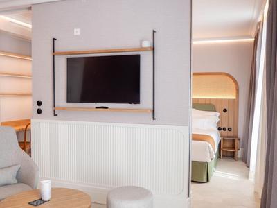 Moderner Hotelzimmerbereich mit Fernseher, grauem Sessel und Blick auf ein Bett im hinteren Raum.