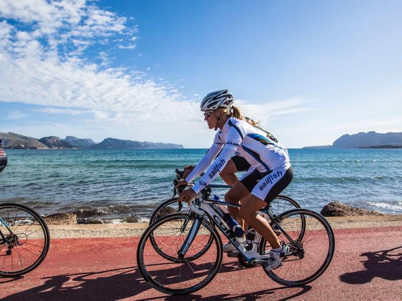 Drei Radfahrer fahren auf einem Weg am Meer bei klarem Himmel und leicht bewölkten Bedingungen.