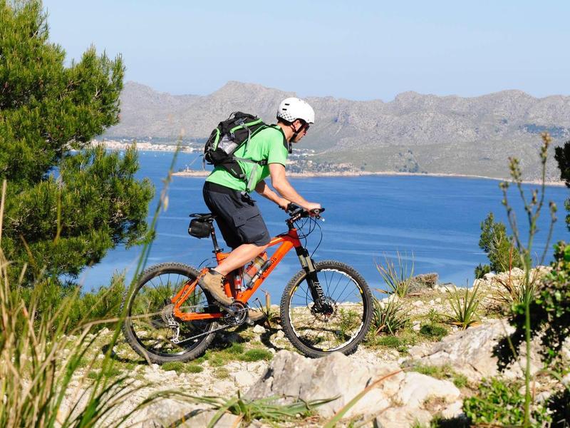 Ein Mann fährt Mountainbike auf felsigem Pfad mit grünem Rucksack, blauem Meer und Bergen im Hintergrund.