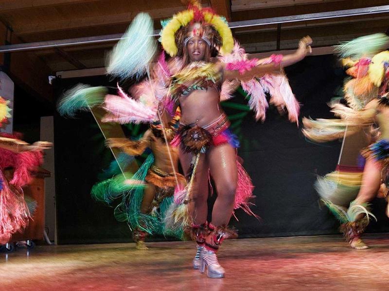 Tänzerinnen in bunten, federverzierten Kostümen tanzen auf einer Bühne.