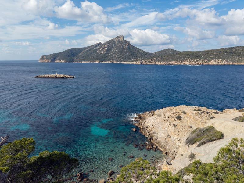 Klares blaues Meer mit felsiger Küste und bewaldeten Hügeln unter blauem Himmel mit Wolken.