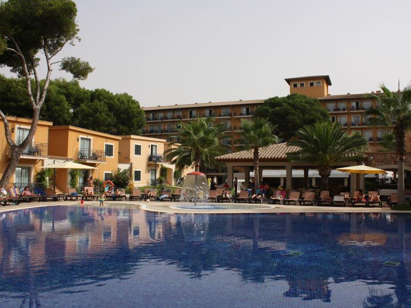 Großer blauer Pool vor gelben Gebäuden mit Palmen und Sonnenschirmen an einem sonnigen Tag.