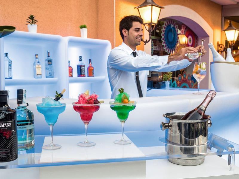 Bar mit weißem Tresen, beleuchtet, Bartender serviert drei farbige Cocktails in bunten Gläsern.