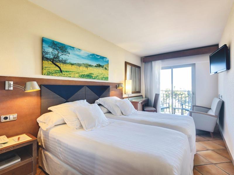 Helles Hotelzimmer mit zwei Einzelbetten, Balkon, Fernseher und Landschaftsgemälde an der Wand.