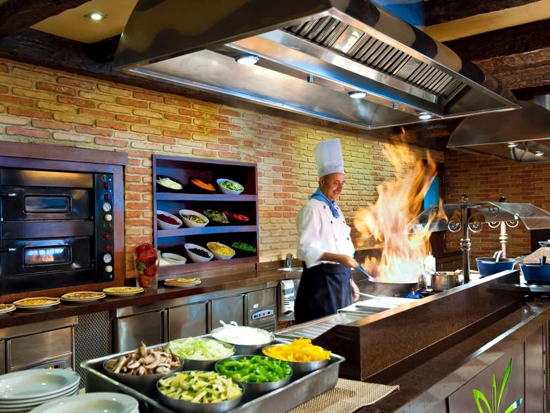 Küche mit zwei Köchen, offener Herd, Flammen, Steinmauern und frischem Gemüse auf der Theke.