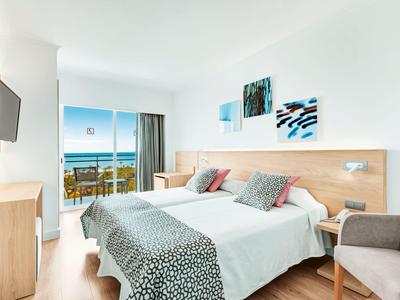 Helles Hotelzimmer mit Doppelbett, Holzmöbeln, Balkon und Meerblick, weiße Wände und moderne Dekoration.