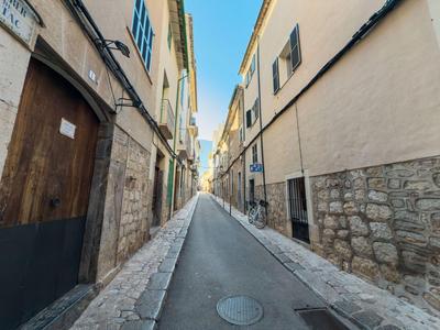 Enge, lange Gasse mit alten Gebäuden aus Stein und Putz, heller Himmel, schmale Gehwege.