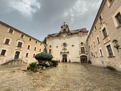 Alter Platz mit barocker Kirche, beige Fassaden, bewölktem Himmel und bepflanztem Brunnen.