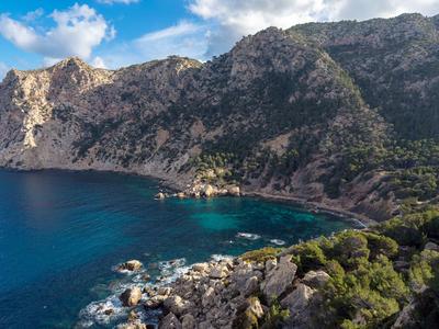Küstenlandschaft mit klarem blauem Wasser, steinigen Felsen und bewaldeten Bergen unter blauem Himmel.