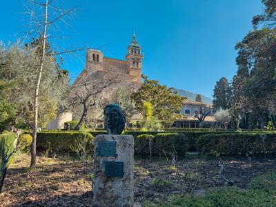 Büste vor gepflegtem Garten, im Hintergrund großes Gebäude mit zwei Türmen und blauem Himmel.
