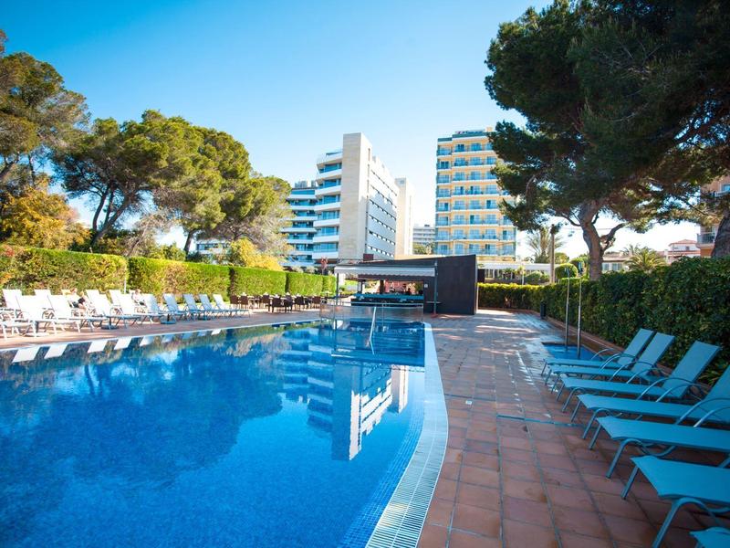 Moderner Außenpool mit blauen Liegestühlen, umgeben von Bäumen und hellen Gebäuden im Hintergrund.