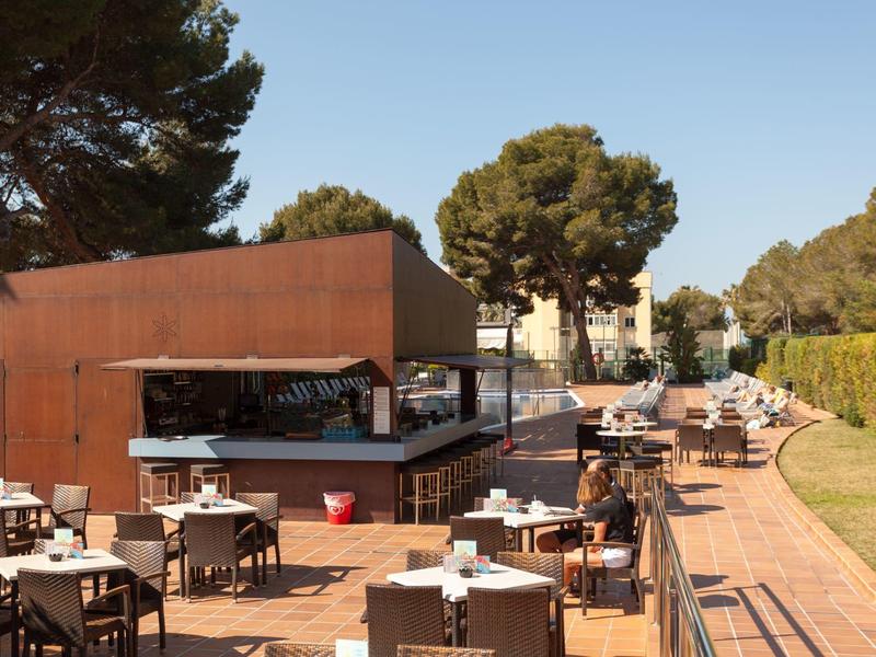 Moderner Außenbereich eines Restaurants mit Holzboden, Tischen, Stühlen und Bäumen im Hintergrund.