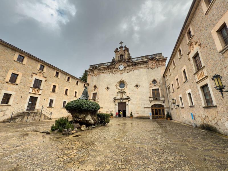 Alter Platz mit barocker Kirche, beige Fassaden, bewölktem Himmel und bepflanztem Brunnen.