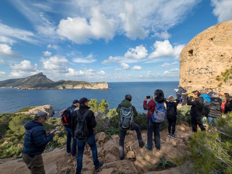Gruppe von Wanderern mit Rucksäcken auf Klippe mit Meer, Himmel mit Wolken und Berg im Hintergrund.