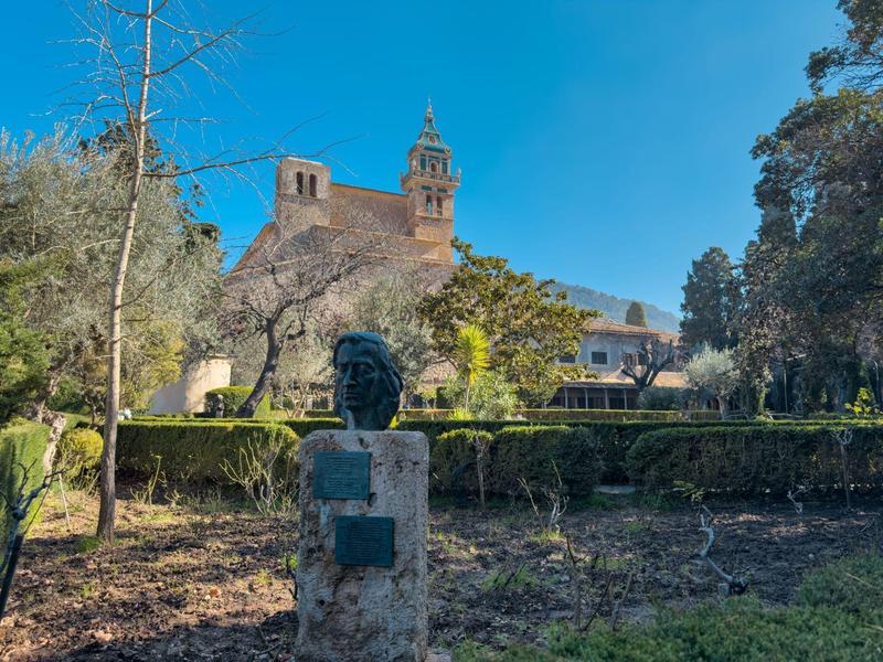Büste vor gepflegtem Garten, im Hintergrund großes Gebäude mit zwei Türmen und blauem Himmel.