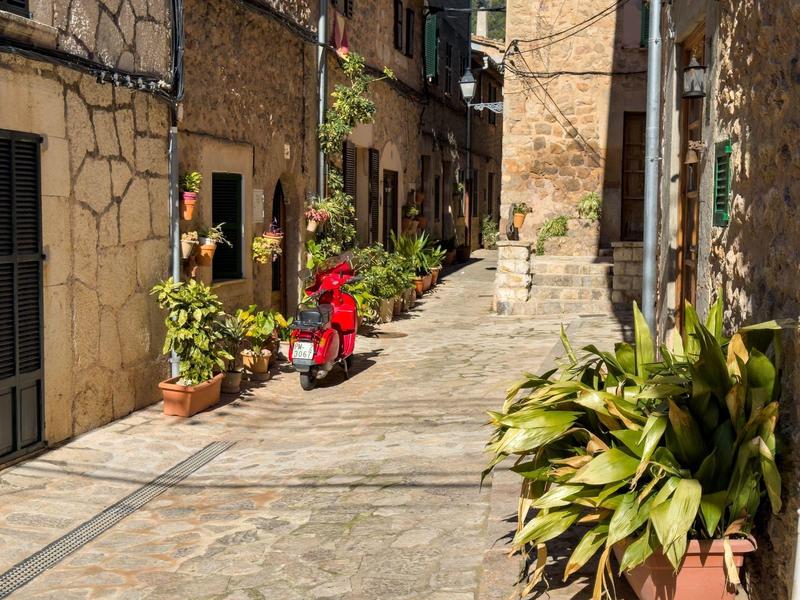 Enge gepflasterte Gasse mit Steinmauern, Pflanzen in Töpfen und rotem Motorroller.