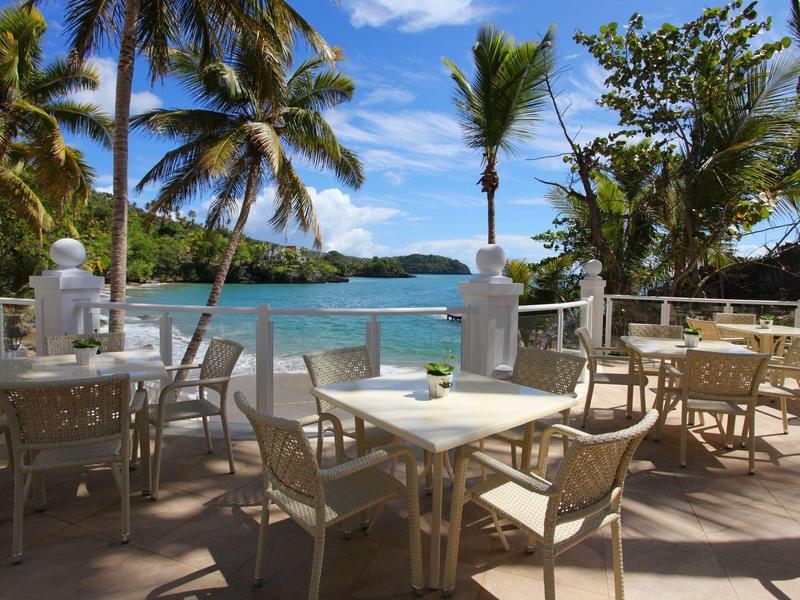 Terrasse mit weißen Tischen und Stühlen, Palmen und Blick auf blaues Meer und bewaldete Inseln.