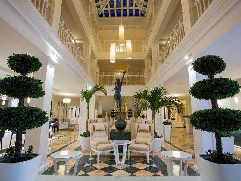 Großer heller Hotellobby mit weißen Möbeln, grünen Pflanzen und einem Kunstwerk in der Mitte.