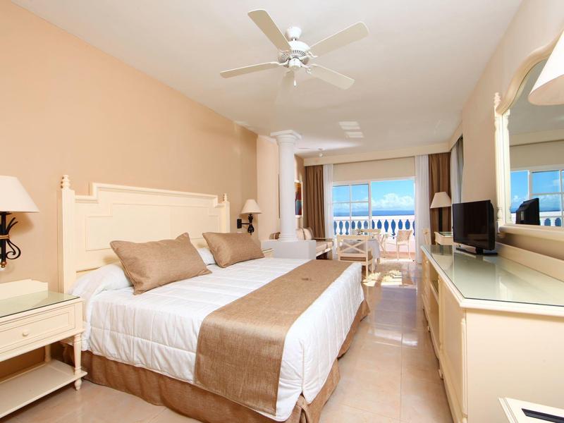 Hell eingerichtetes Schlafzimmer mit Doppelbett, Ventilator, Fernseher und Balkon mit Meerblick.