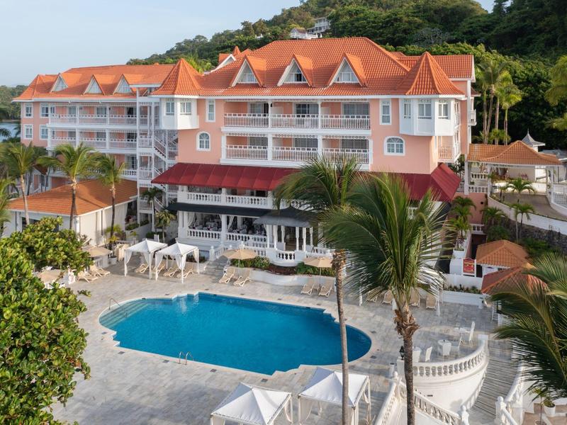 Großes Hotel mit orangem Dach, weißem Balkon, umgeben von Palmen und großem blauem Pool.