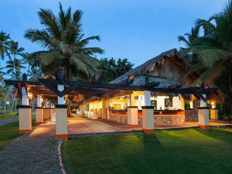 Ein tropisches, offenes Restaurant mit hellen Säulen, Holzdach und Palmen im Hintergrund bei Abendlicht.