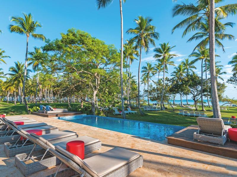 Ein Pool mit Liegestühlen und Palmen, blauer Himmel und Meer im Hintergrund.