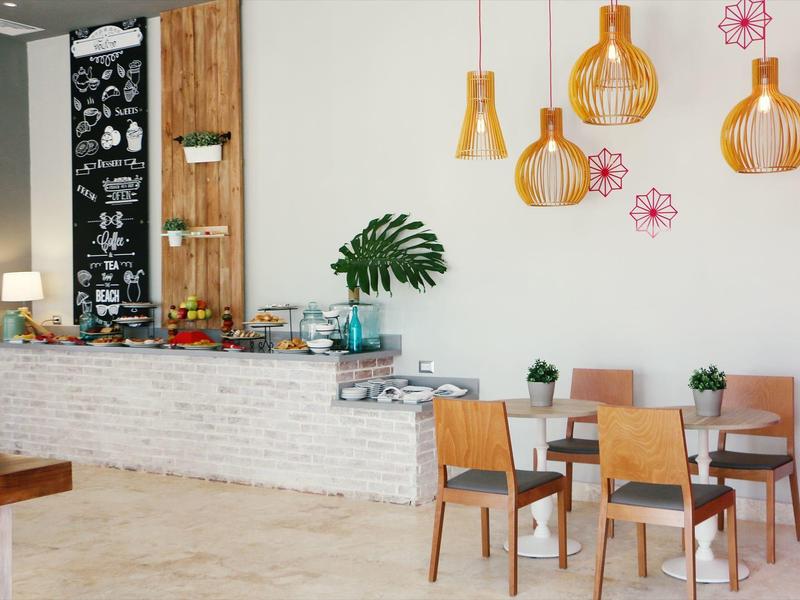 Heller Gastraum mit Holzstühlen, weißen Tischen, Hängelampen und Pflanzen an heller Wand.