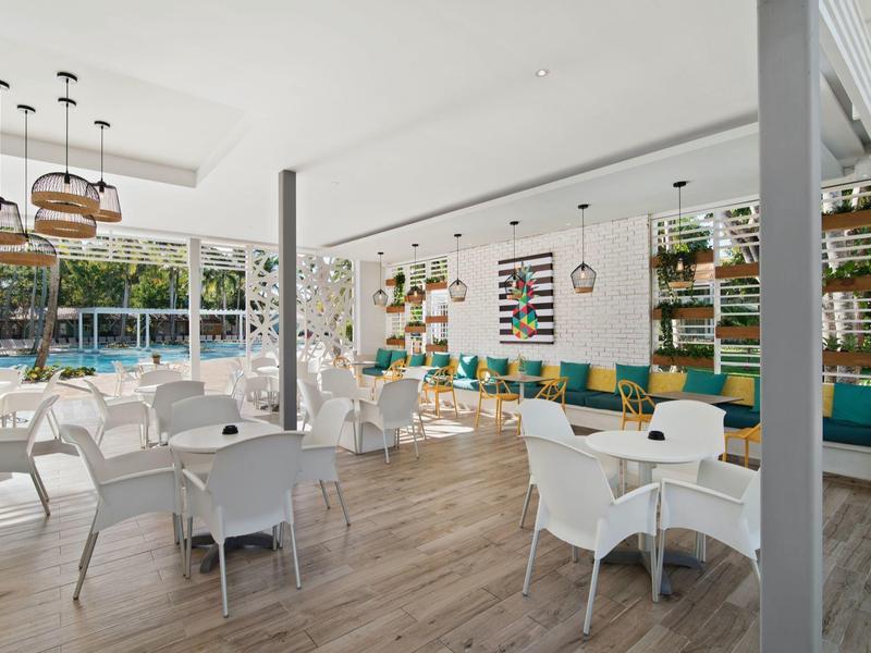 Moderner Cafébereich mit weißen Stühlen, Holzboden und großen Fenstern, viel Tageslicht.