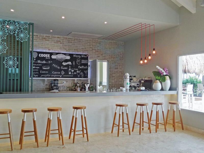 Moderne Cafébar mit weißem Tresen, Holzhockern, Hängelampen und großer Fensterfront.