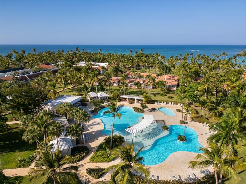 Großer Außenpool mit Sonnenschirmen, Palmen und Blick auf das Meer im Hintergrund.
