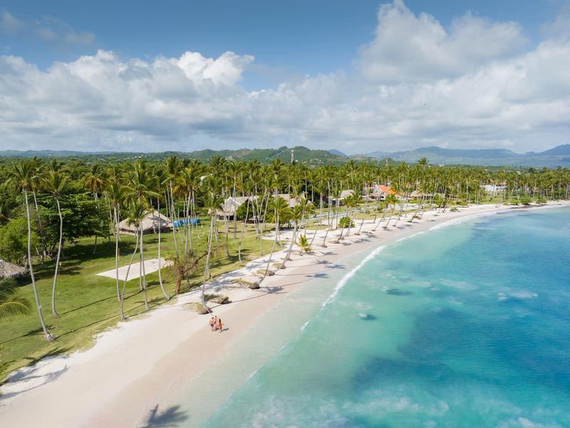 Ein weißer Sandstrand mit Palmen, türkisblauem Meer und teilweise bewölktem Himmel.
