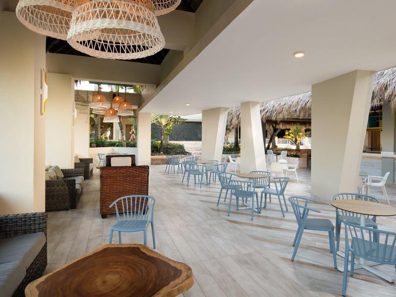 Moderner, heller Außenbereich mit blauen Stühlen, Holztischen und geflochtenen Lampen an der Decke.