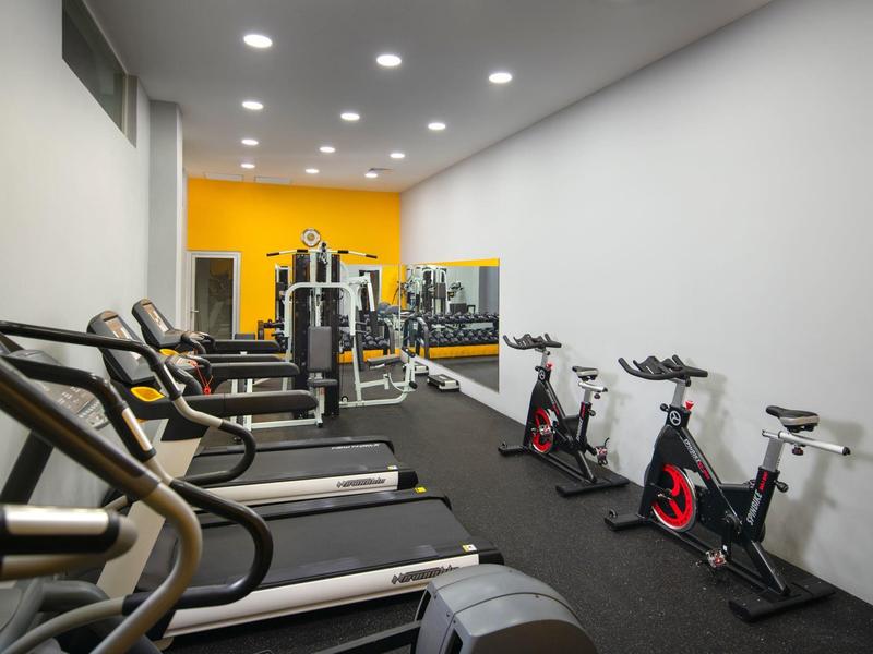 Moderner Fitnessraum mit Laufbändern links, stationären Fahrrädern rechts, Spiegelrückwand und gelber Akzentwand.