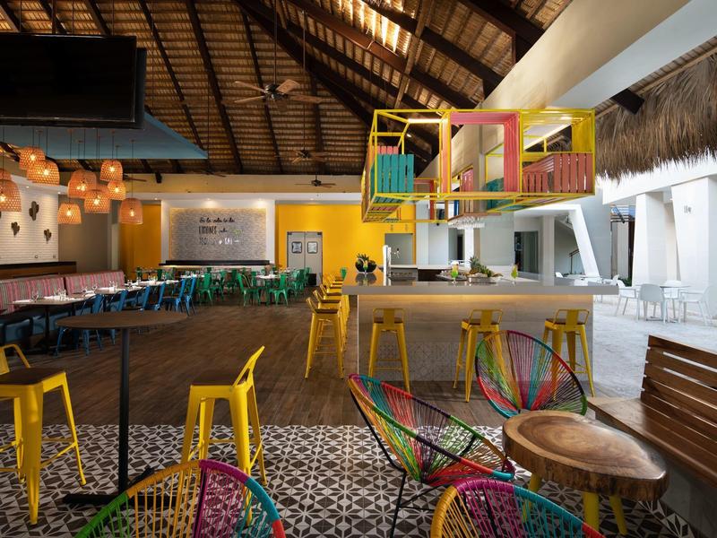 Offener, farbenfroher Gastraum mit gelben Stühlen, bunten Sitzkörben und gedecktem Holzdekor.