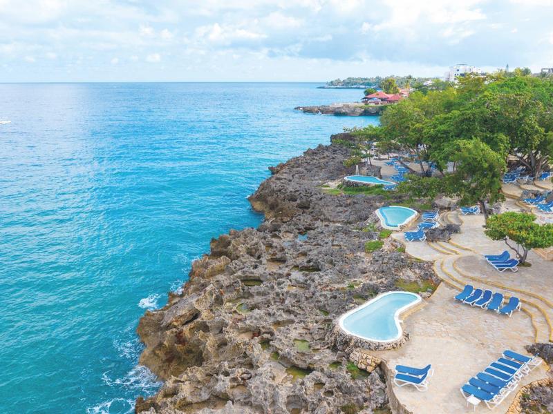 Klippenküste mit blauem Meer, mehreren kleinen Pools und Liegestühlen unter Bäumen.