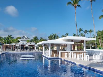 Großer blauer Pool mit weißer Bar und Palmen unter blauem Himmel in einem Resort.