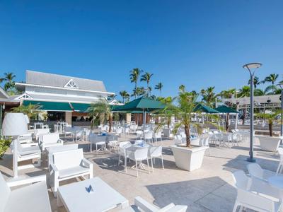 Moderner Außenbereich eines Restaurants mit weißen Tischen, Stühlen, Sonnenschirmen und Palmen bei blauem Himmel.