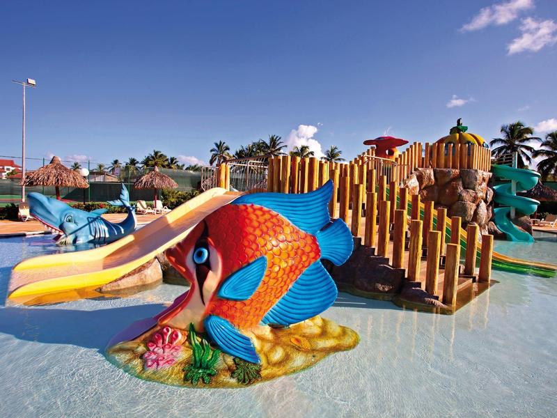 Bunte Wasserrutsche mit Fischfigur und Holzbrücke in flachem Wasser bei klarem Himmel.