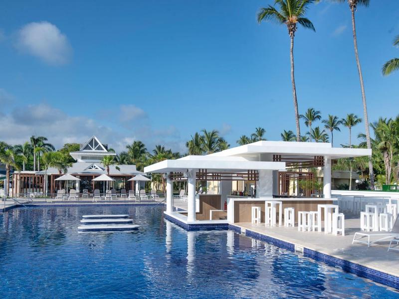 Großer blauer Pool mit weißer Bar und Palmen unter blauem Himmel in einem Resort.