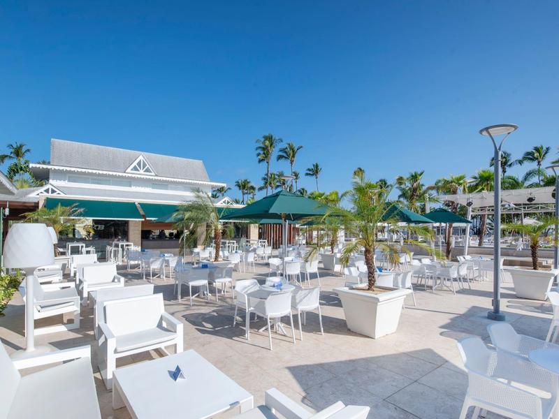 Moderner Außenbereich eines Restaurants mit weißen Tischen, Stühlen, Sonnenschirmen und Palmen bei blauem Himmel.