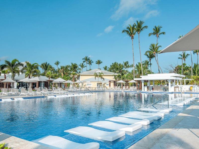 Großer Pool mit weißen Liegen und Palmen unter klarem blauem Himmel in einem Resort.