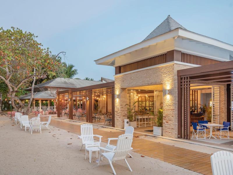 Ein helles Strandrestaurant mit weißen Stühlen, Bäumen und blassem blauen Himmel im Hintergrund.