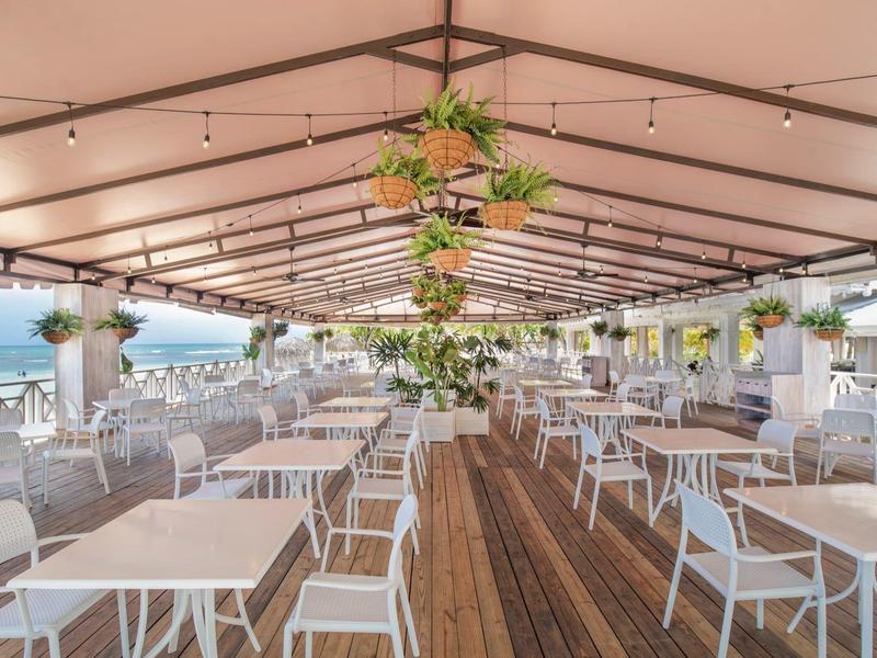 Hell überdachte Terrasse mit weißen Tischen, Stühlen, Holzfußboden und hängenden Pflanzen nahe Meer.
