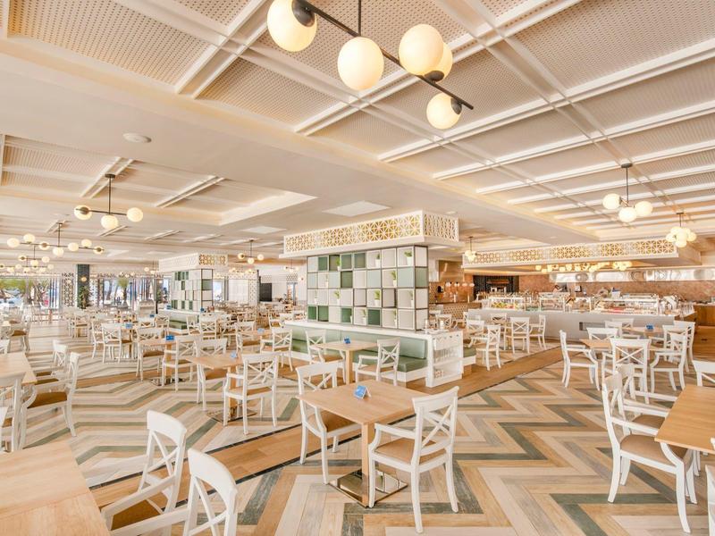 Weites, modernes Restaurant mit hellen Holzmöbeln, geometrischem Bodenmuster und Deckenleuchten.