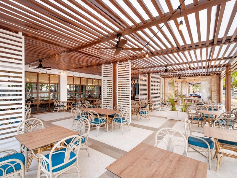 Offener Gastronomiebereich mit Holzdach, weißen Stühlen, Holztischen und blau gepolsterten Sitzkissen.