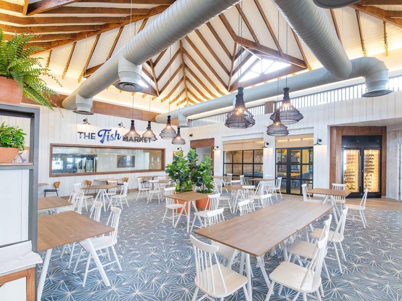 Helle, moderne Restaurant mit weißen Stühlen, Holztischen, Pflanzen und sichtbaren Lüftungsrohren.