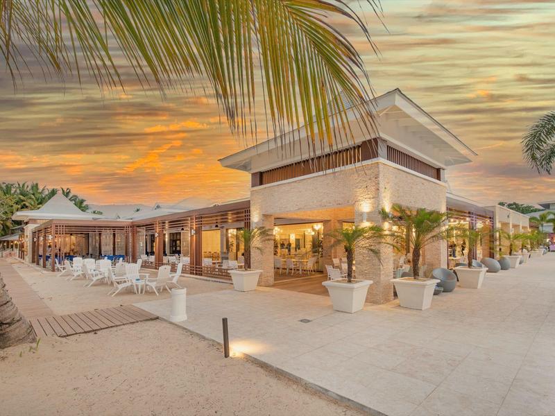 Moderner Strandclub mit Palmen, Sandboden und orangefarbenem Sonnenuntergang im Hintergrund.