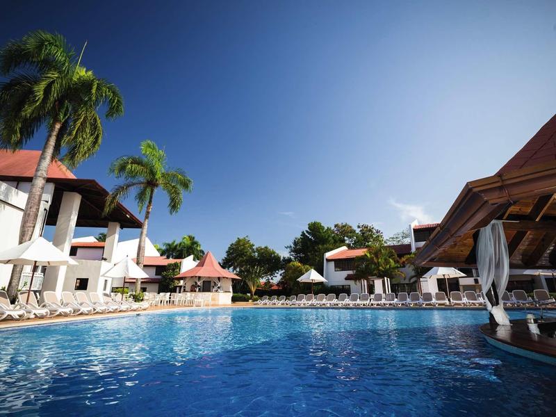 Klares Schwimmbecken vor weißen Gebäuden mit roten Dächern und Palmen unter blauem Himmel.