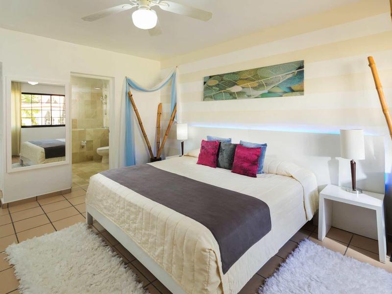 Helles Schlafzimmer mit großem Bett, bunten Kissen, Teppich und moderner Wanddekoration.